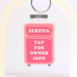 May include: A pink luggage-shaped keychain with the name "SERENA" and the text "TAP FOR OWNER INFO" in white. The keychain has a silver ring and a gray handle. The background is a light cream color.