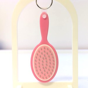 Peut inclure: Un porte-cl&eacute;s rose en forme d'ovale repr&eacute;sentant une mini brosse &agrave; cheveux. La brosse a un manche rose et un bord blanc autour de la t&ecirc;te ovale, remplie de petites soies argent&eacute;es. Le porte-cl&eacute;s est suspendu &agrave; un anneau argent&eacute;.