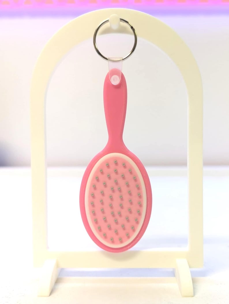 Peut inclure: Un porte-cl&eacute;s rose en forme d'ovale repr&eacute;sentant une mini brosse &agrave; cheveux. La brosse a un manche rose et un bord blanc autour de la t&ecirc;te ovale, remplie de petites soies argent&eacute;es. Le porte-cl&eacute;s est suspendu &agrave; un anneau argent&eacute;.