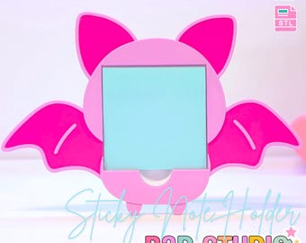 May include: A pink bat-shaped sticky note holder with a light blue note inside. The holder has a square opening for the note and wings on either side. Text on the image reads "3D PRINT FILE" and "Sticky Note Holder".