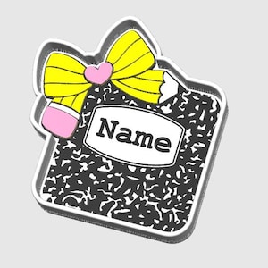 May include: A sticker featuring a composition notebook design with a yellow bow, a pink heart, and a pencil. The notebook has a white label with the word "Name" in black. The background is a light gray.