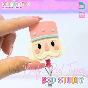 May include: A pink and cream-colored badge reel topper in the shape of a nutcracker soldier. The topper has a pink hat with blue accents and a white mustache. The text "3D PRINT FILE" and "Badge Reel Topper" are visible.