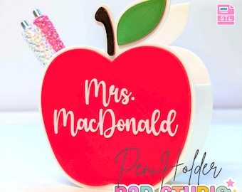 Könnte beinhalten: Ein roter, apfelförmiger Stifthalter mit dem Namen "Mrs. MacDonald" in weißer Schrift. Der Stifthalter hat ein grünes Blatt und einen braunen Stiel. Die Wörter "Pen Holder" und "B3D STUDIO" sind ebenfalls sichtbar. Zwei Glitzerstifte befinden sich darin.