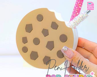 May include: A cookie-shaped pen holder with a bite taken out of it, featuring brown chocolate chip details. Two glitter pens are inside. The text "3D PRINT FILE" and "Pen Holder B3D STUDIO" are visible.