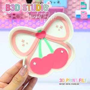 May include: A white and pink trinket dish in the shape of cherries and a bow. The dish has a pink bow with a heart in the center and two pink cherries with white diamond accents. The text "B3D STUDIO" and "Trinket Dish" are at the top.