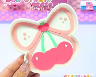 May include: A white and pink trinket dish in the shape of cherries and a bow. The dish has a pink bow with a heart in the center and two pink cherries with white diamond accents. The text "B3D STUDIO" and "Trinket Dish" are at the top.