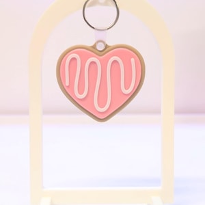 May include: A pink heart-shaped keychain with white icing detail, hanging from a silver ring. The keychain is set against a cream-colored arch-shaped display stand with a rectangular base. The design resembles a cookie.