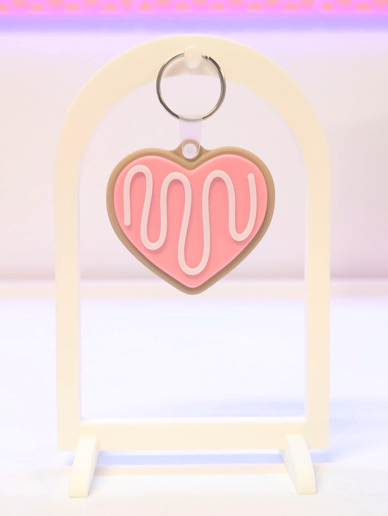 May include: A pink heart-shaped keychain with white icing detail, hanging from a silver ring. The keychain is set against a cream-colored arch-shaped display stand with a rectangular base. The design resembles a cookie.