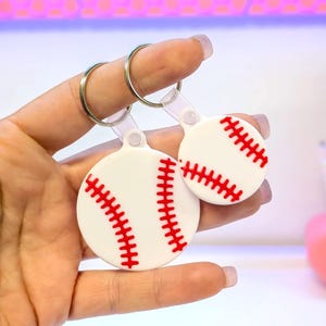 May include: Two baseball-themed keyrings. Each keyring features a white, circular design with red stitching, resembling a baseball. The keyrings are attached to silver rings.
