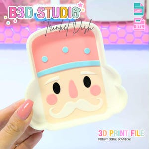 May include: A whimsical trinket dish in the shape of a nutcracker, featuring a pink and blue hat, a cream-colored face, and a white mustache. The dish has a rectangular shape with rounded edges. Text on the image reads "B3D STUDIO Trinket Dish" and "3D PRINT FILE".