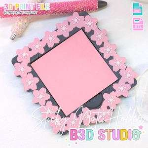 May include: A pink sticky note holder with a gray frame, adorned with white cherry blossom accents. The holder contains a pink sticky note pad. The text "3D PRINT FILE" and "Sticky Note Holder" are visible.