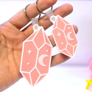 May include: Two pink crystal-shaped keychains with white outlines, each featuring a crescent moon and stars. The keychains are attached to silver key rings. The background is a gradient of pink and purple.
