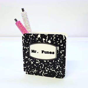 May include: A small, white pen holder with a black and white composition notebook design. The pen holder has the text "Mr. Tunes" on the front. Two pens with rhinestone embellishments, one pink and one white, are inside the holder.