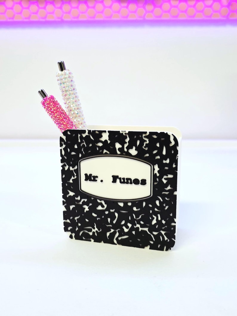 May include: A small, white pen holder with a black and white composition notebook design. The pen holder has the text "Mr. Tunes" on the front. Two pens with rhinestone embellishments, one pink and one white, are inside the holder.