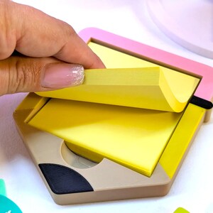 May include: A pencil-shaped memo pad holder with a pink and yellow frame. The holder is beige with a black accent and contains a stack of yellow sticky notes. A hand is lifting a note from the pad.