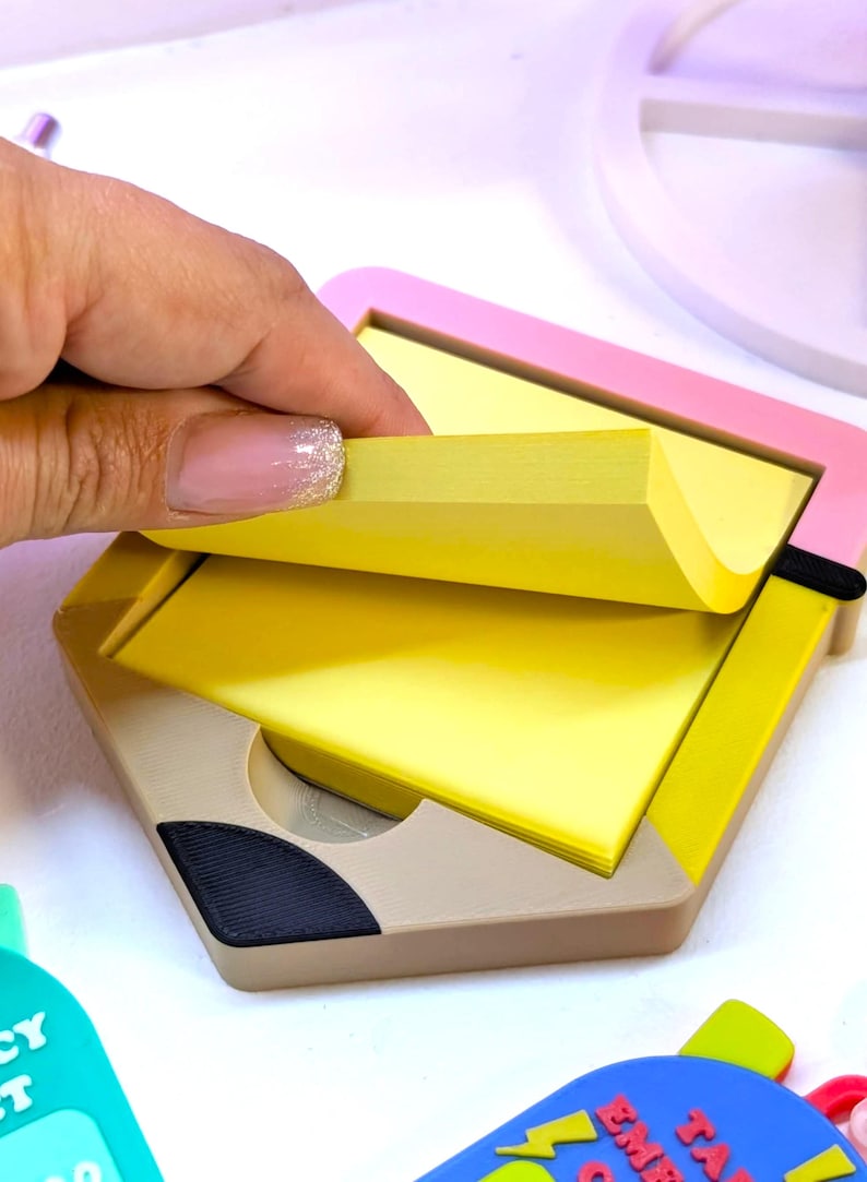 May include: A pencil-shaped memo pad holder with a pink and yellow frame. The holder is beige with a black accent and contains a stack of yellow sticky notes. A hand is lifting a note from the pad.