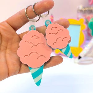 May include: Two pink cotton candy-shaped keychains with blue and white striped cone bottoms. The keychains are held up by a silver ring and a clear plastic strap. The background is blurred, with hints of other colorful items.