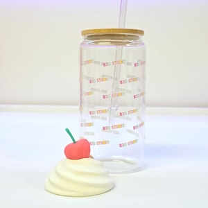 May include: A clear glass tumbler with a bamboo lid and a clear straw. The tumbler is printed with the text "B3D STUDIO" in a repeating pattern. A decorative cream-colored swirl with a red cherry on top sits in front of the tumbler.