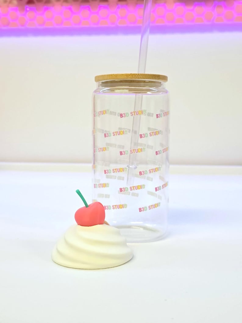 May include: A clear glass tumbler with a bamboo lid and a clear straw. The tumbler is printed with the text "B3D STUDIO" in a repeating pattern. A decorative cream-colored swirl with a red cherry on top sits in front of the tumbler.