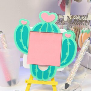May include: A whimsical cactus-shaped memo board with a pink square for notes, set on a yellow easel. The cactus is teal with pink heart accents. A sparkly pen and cactus keychains are also visible.