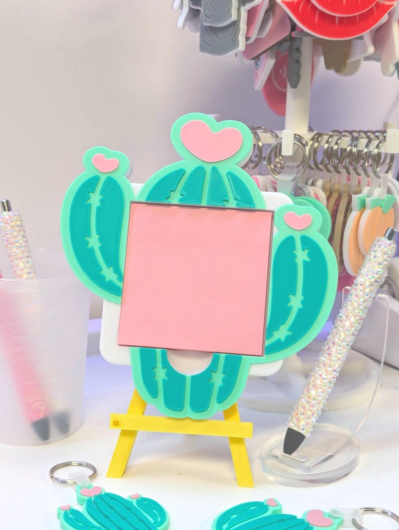 May include: A whimsical cactus-shaped memo board with a pink square for notes, set on a yellow easel. The cactus is teal with pink heart accents. A sparkly pen and cactus keychains are also visible.