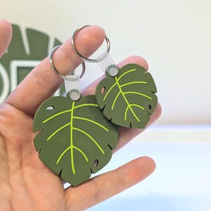 Puede incluir: Dos llaveros con forma de hoja. Los llaveros son de color verde oliva con detalles de venas verdes brillantes. Cada llavero tiene un anillo de metal plateado y una etiqueta de plástico transparente. Los llaveros se sostienen en una mano sobre un fondo blanco.