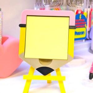 May include: A yellow pencil-shaped desk organizer with a pink top and a yellow easel base. The organizer has a yellow notepad and a small drawer. The design includes black and tan accents. The word "Mason" is visible on a sticker.