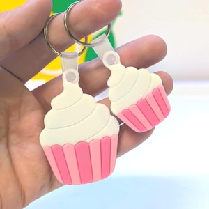 May include: Two cupcake-shaped keychains. Each keychain features a white frosting swirl atop a pink cupcake liner. The keychains are attached to silver key rings. The cupcakes are made of a soft, rubbery material.
