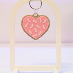 May include: A heart-shaped keychain resembling a pink frosted cookie with white sprinkles and a brown border. The keychain hangs from a silver ring and is displayed on a white stand.