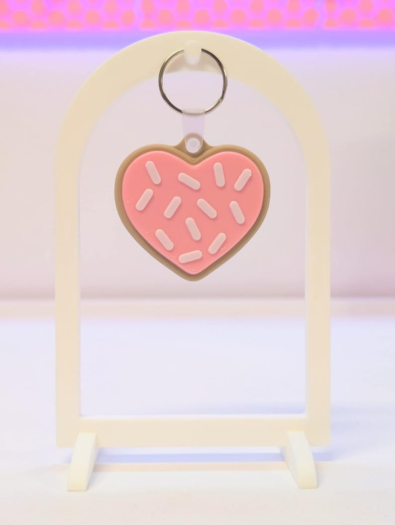 May include: A heart-shaped keychain resembling a pink frosted cookie with white sprinkles and a brown border. The keychain hangs from a silver ring and is displayed on a white stand.