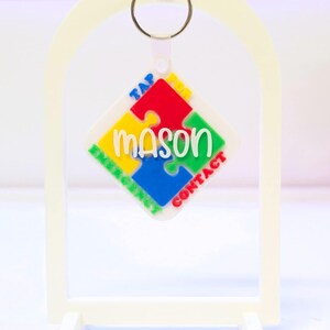 May include: A white keychain with a puzzle piece design. The puzzle piece is made up of red, yellow, green, and blue sections, with the name "mason" in green. The keychain also has the words "TAP EMERGENCY CONTACT" in colorful text.