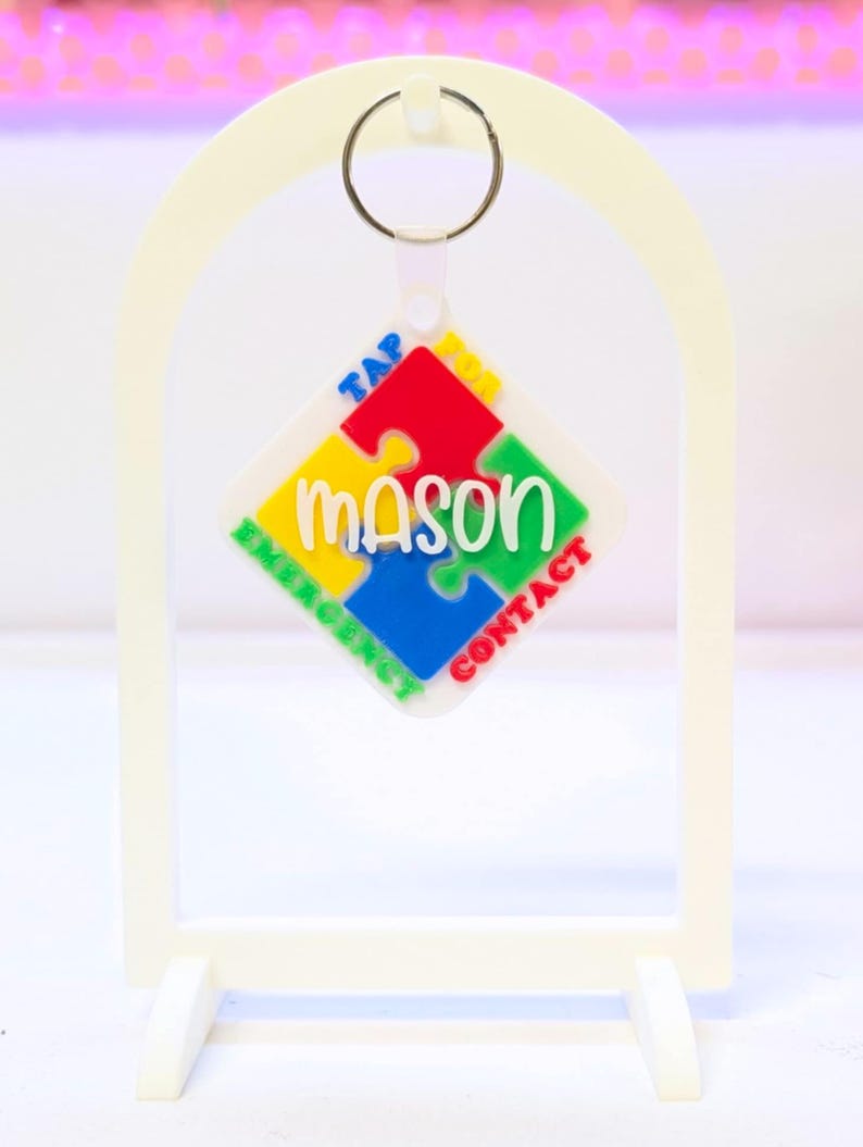 May include: A white keychain with a puzzle piece design. The puzzle piece is made up of red, yellow, green, and blue sections, with the name "mason" in green. The keychain also has the words "TAP EMERGENCY CONTACT" in colorful text.