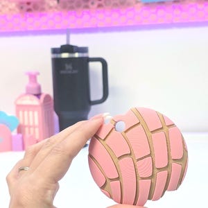 Puede incluir: Un objeto rosa y beige en forma de concha con un agujero en la parte superior. El objeto se sostiene en la mano de una persona. El fondo incluye una taza negra, estantes rosas y otros objetos rosas.