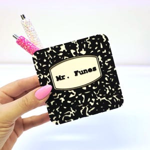 May include: A black and white composition notebook-style coaster with the name "Mr. Funes" printed on it. Two decorative pens with glitter accents are in the background. The coaster is held by a hand with pink nail polish.