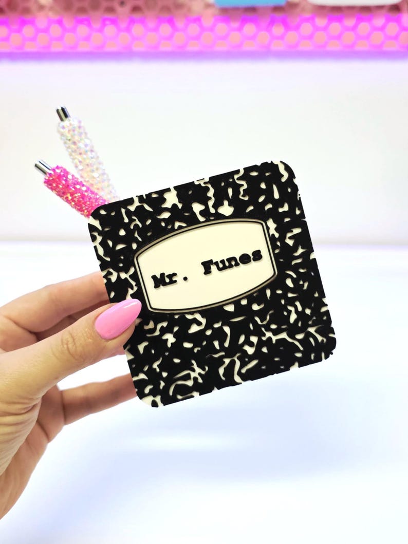 May include: A black and white composition notebook-style coaster with the name "Mr. Funes" printed on it. Two decorative pens with glitter accents are in the background. The coaster is held by a hand with pink nail polish.