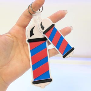 Puede incluir: Un llavero de poste de barbería con la parte superior e inferior blancas, rayas rojas y azules y una banda negra. El llavero está sujeto a un llavero plateado.