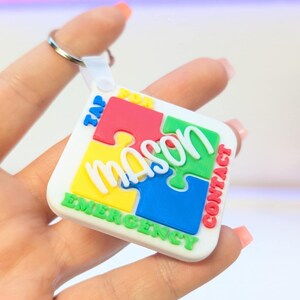 May include: A white puzzle-shaped keychain with the name "Mason" in green, yellow, red, and blue puzzle pieces. The keychain has the words "TAP FOR CONTACT EMERGENCY" in white and red text. The keychain is held by a person.