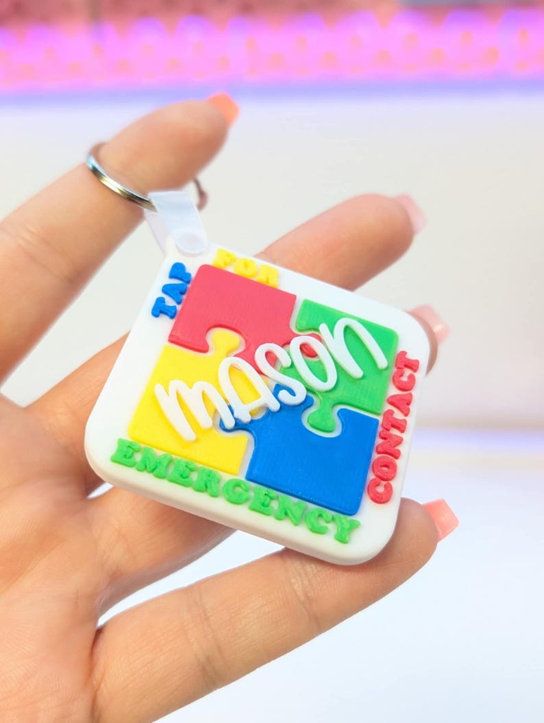 May include: A white puzzle-shaped keychain with the name "Mason" in green, yellow, red, and blue puzzle pieces. The keychain has the words "TAP FOR CONTACT EMERGENCY" in white and red text. The keychain is held by a person.