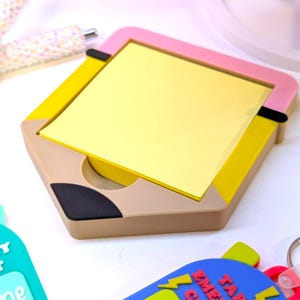 May include: A hexagonal pencil-shaped notepad holder with a pink frame, yellow sides, and a beige base. The notepad is filled with yellow sticky notes. Also visible are a pen and two keychains with text.