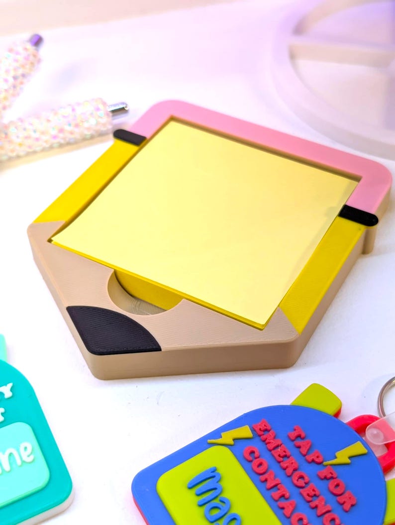 May include: A hexagonal pencil-shaped notepad holder with a pink frame, yellow sides, and a beige base. The notepad is filled with yellow sticky notes. Also visible are a pen and two keychains with text.