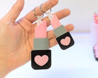 May include: Two lipstick-shaped keychains. The lipstick has a pink bullet, a gray midsection, and a black base with a pink heart. The keychains are held by a silver ring and a clear plastic connector.