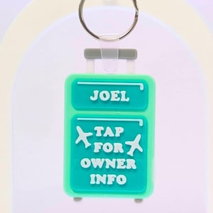 Op de afbeelding: Een turquoise bagagelabelsleutelhanger met de naam "JOEL" bovenaan. Daaronder staat de tekst "TAP FOR OWNER INFO" met vliegtuigafbeeldingen. De sleutelhanger heeft een zilveren ring en een grijze handgreep.