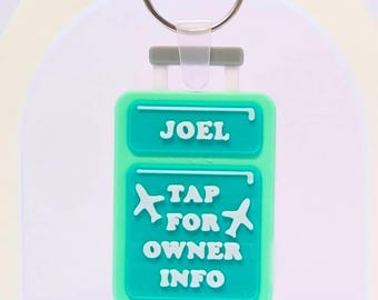 Puede incluir: Llavero de etiqueta de equipaje turquesa con el nombre "JOEL" en la parte superior. Debajo, el texto dice "TAP FOR OWNER INFO" con gráficos de avión. El llavero tiene un anillo plateado y un asa gris.