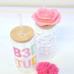 May include: Two clear glass tumblers with bamboo lids and clear straws. One tumbler has the text "B3D STUDIO" in colorful letters. The other has a pink rose lid. A small pink rose decoration is also present.
