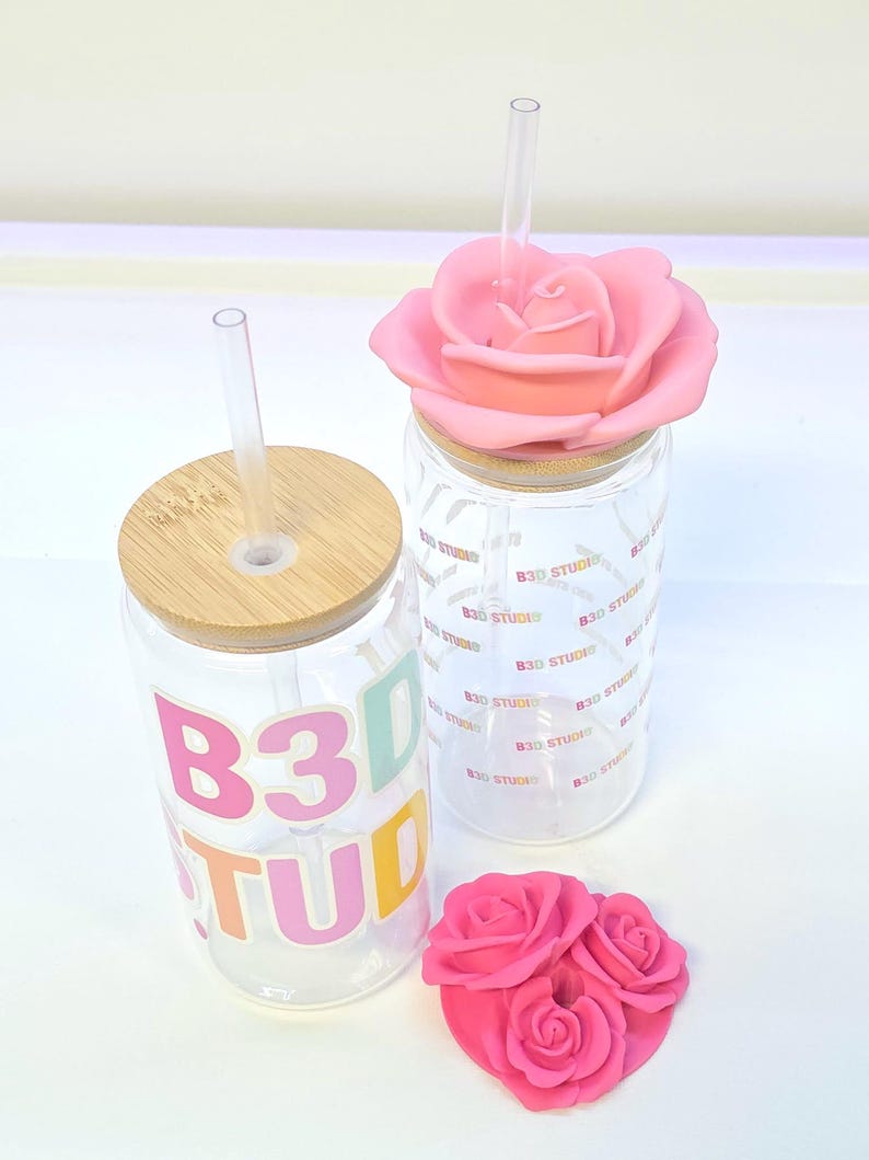 May include: Two clear glass tumblers with bamboo lids and clear straws. One tumbler has the text "B3D STUDIO" in colorful letters. The other has a pink rose lid. A small pink rose decoration is also present.
