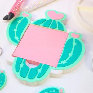 May include: A cactus-shaped memo holder with a pink sticky note, teal green cactus details, and a pink heart accent. Two glitter pens in pink and white are in the background. A keychain with a cactus design is also visible.