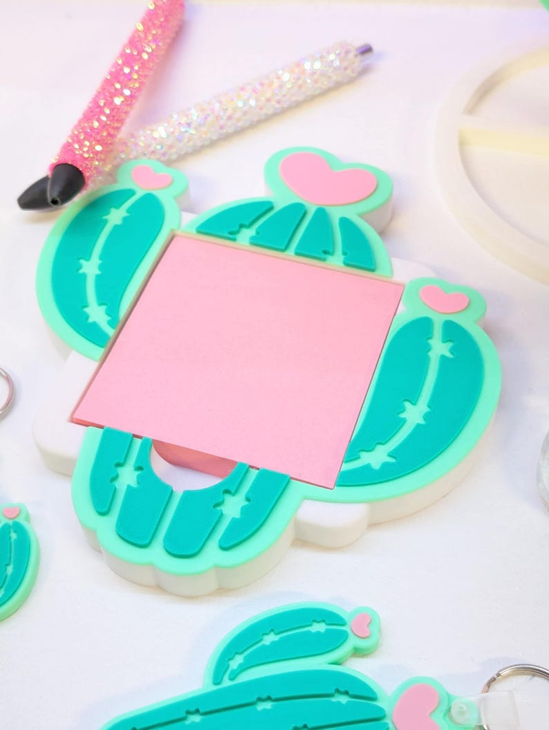 May include: A cactus-shaped memo holder with a pink sticky note, teal green cactus details, and a pink heart accent. Two glitter pens in pink and white are in the background. A keychain with a cactus design is also visible.