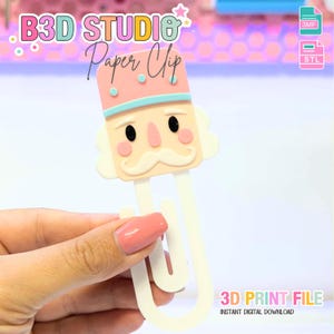 May include: A novelty paper clip in the shape of a nutcracker soldier. The clip is white with a pink, blue, and beige nutcracker head. The image includes the text "B3D STUDIO Paper Clip" and "3D PRINT FILE INSTANT DIGITAL DOWNLOAD".