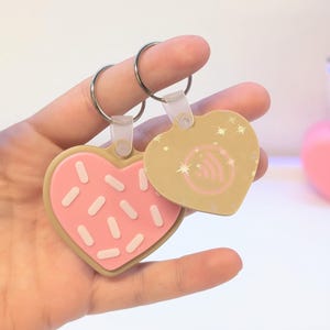 May include: Two heart-shaped keychains. One is designed to look like a pink frosted cookie with white sprinkles. The other is tan with a wireless symbol and star accents. Both have silver key rings.