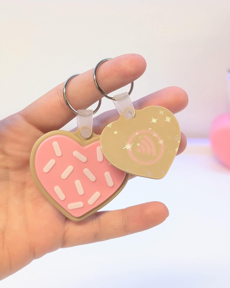May include: Two heart-shaped keychains. One is designed to look like a pink frosted cookie with white sprinkles. The other is tan with a wireless symbol and star accents. Both have silver key rings.